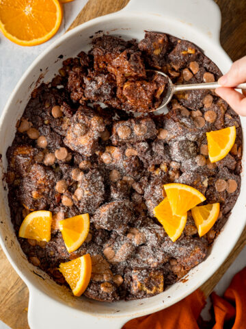 Overhead square image of chocolate orange bread and butter pudding in a white dish, set on a wooden board. The pudding has orange slices on top and a spoonful is being taken.
