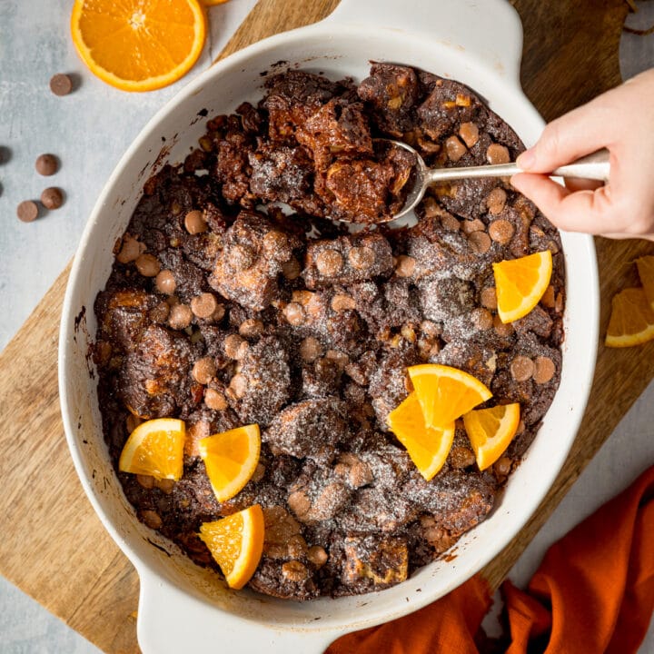 Overhead square image of chocolate orange bread and butter pudding in a white dish, set on a wooden board. The pudding has orange slices on top and a spoonful is being taken.