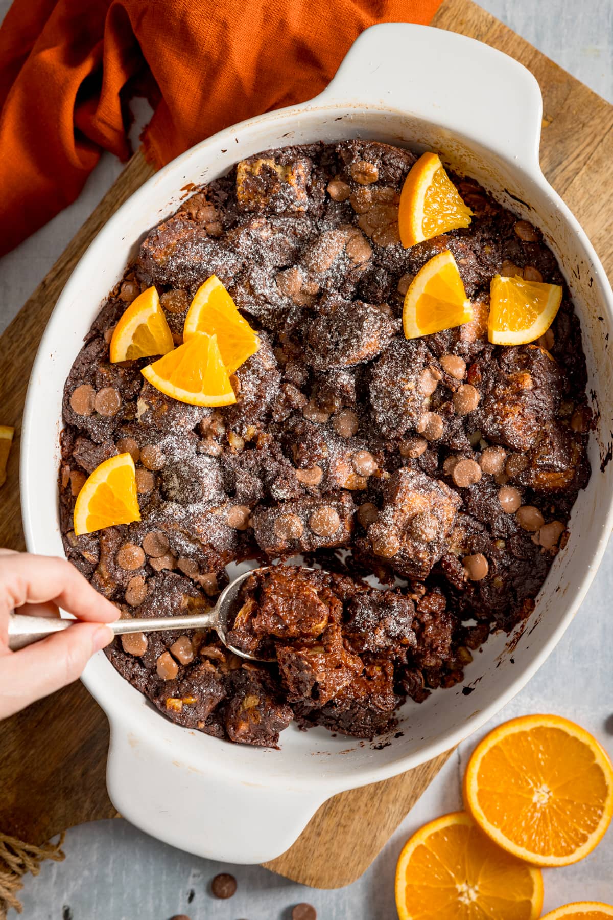 Overhead image of chocolate orange bread and butter pudding in a white dish, set on a wooden board. The pudding has orange slices on top and a spoonful is being taken. There are orange slices and an orange napkin around the dish.