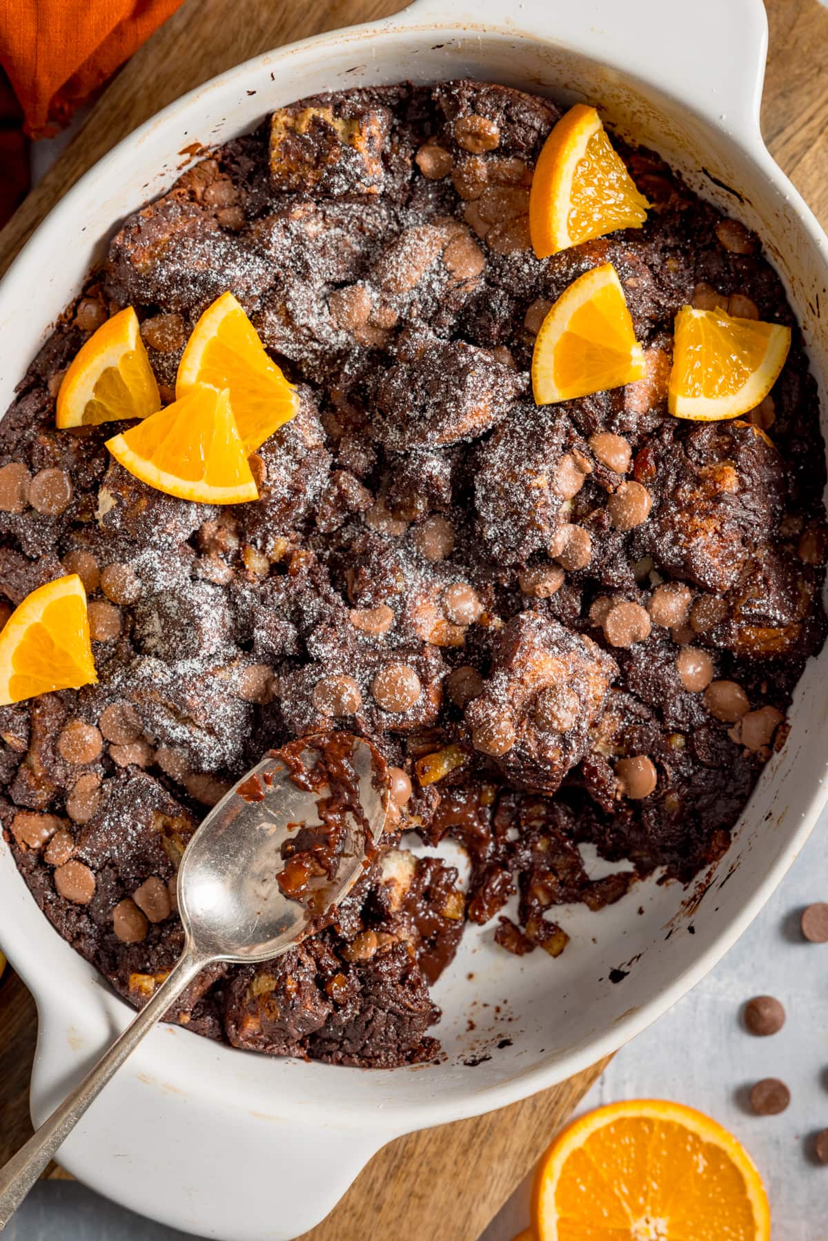 Tall image of chocolate orange bread and butter pudding in a white dish, set on a wooden board. The pudding has orange slices on top and a spoonful is being taken.