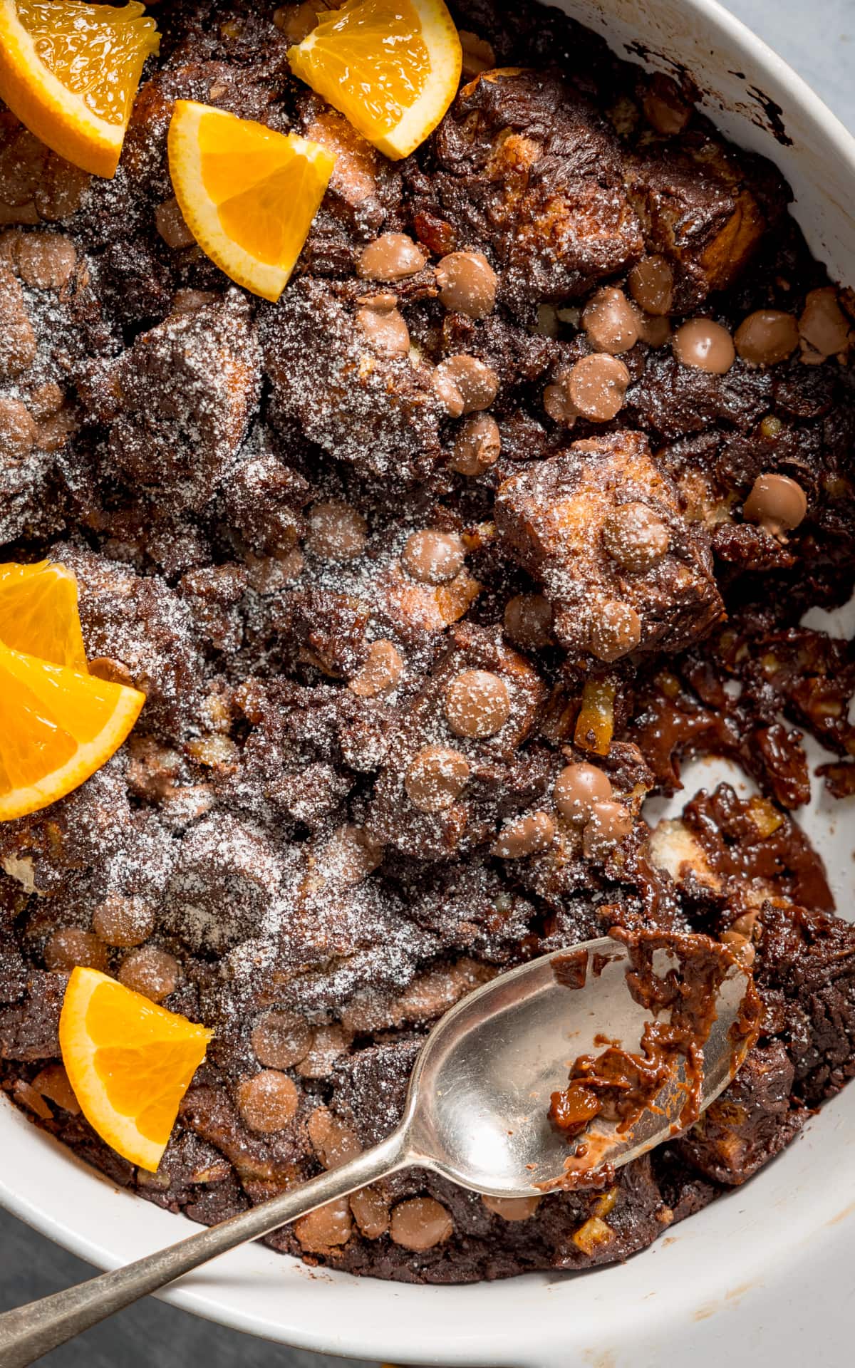Close up overhead image of chocolate orange bread and butter pudding in a white dish. The pudding has orange slices on top and a spoonful is being taken.
