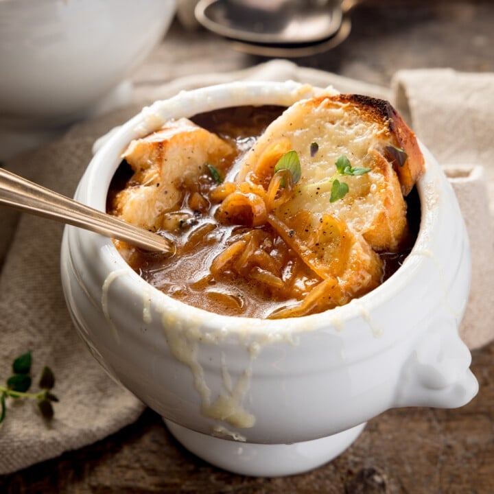 Close-up square image of French Onion Soup in a white bowl on a wooden table next to a beige napkin. There is a gold spoon sticking out of the bowl.