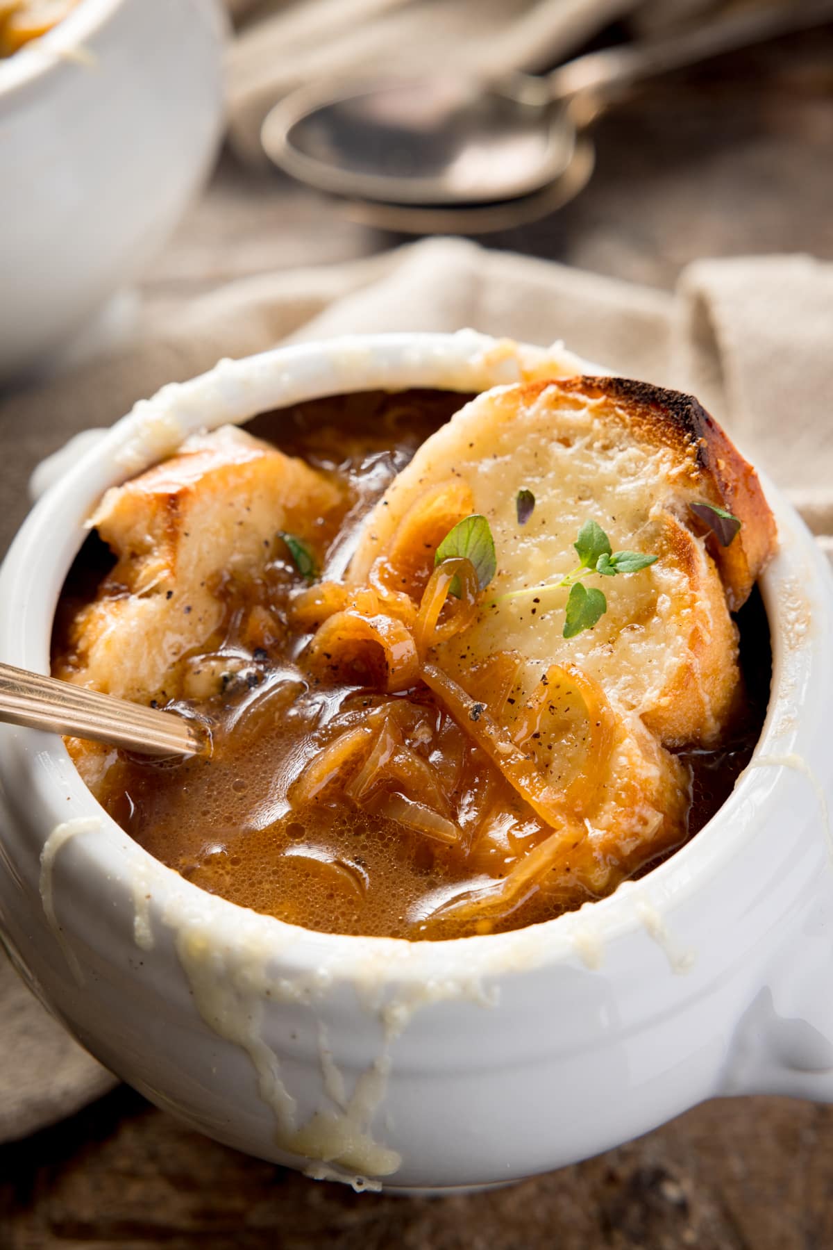 Close-up of French Onion Soup in a white bowl on a wooden table next to a beige napkin. There is a gold spoon sticking out of the bowl. There is a further bowl and two spoons just in shot at the top of the image.