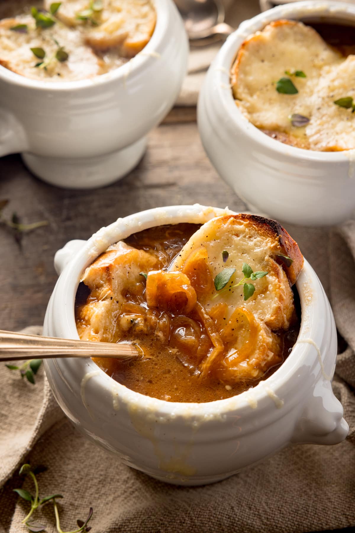 Overhead shot of three bowls of French Onion Soup in white bowls. The bowls are on a wooden table next to a beige napkin. There is a gold spoon sticking out of the bowl at the front of the image.