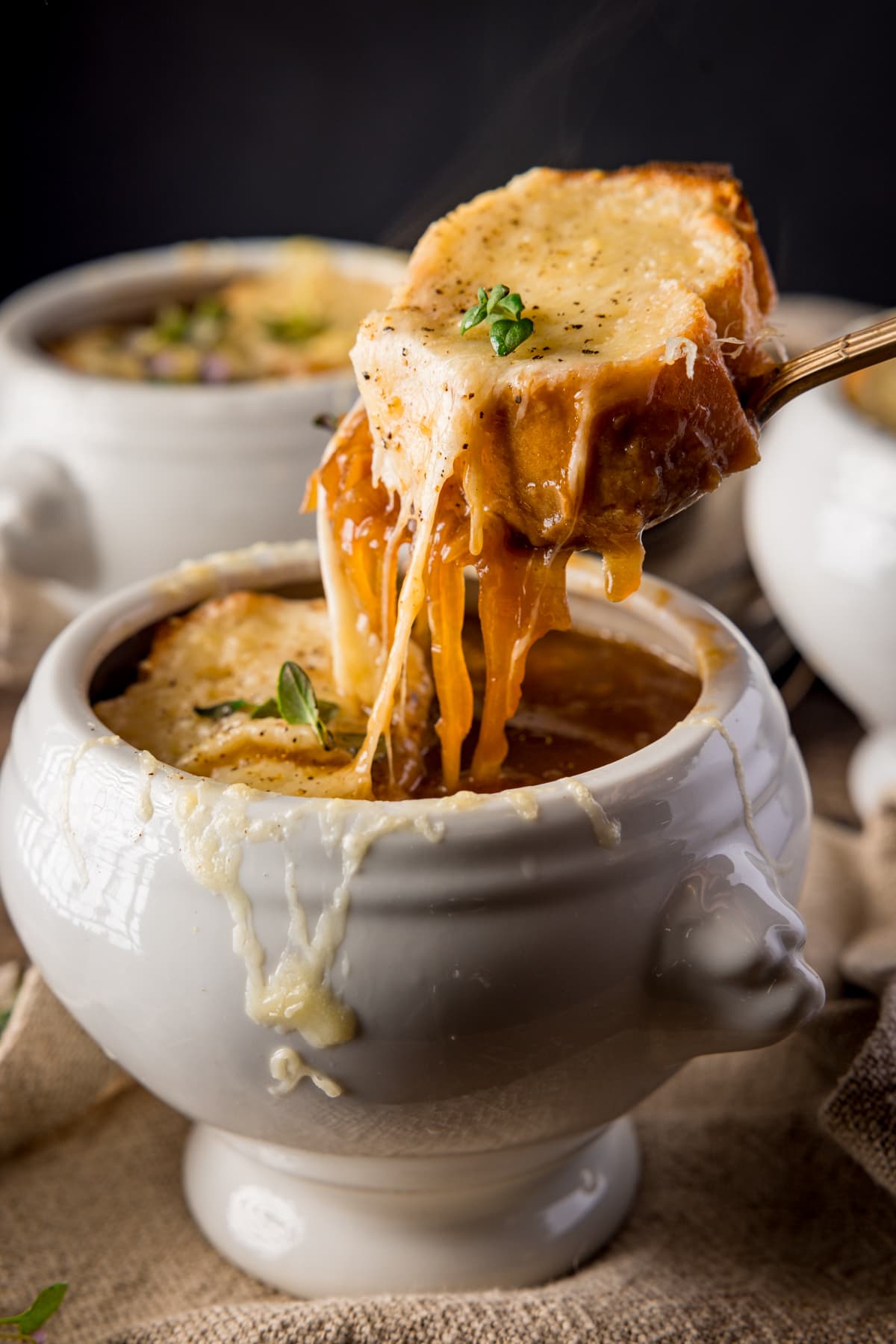 A spoonful of cheese-topped-baguette-slice being taken from a bowl of French Onion Soup. The soup is in a white bowl on a wooden table. There are two further bowls in the background.