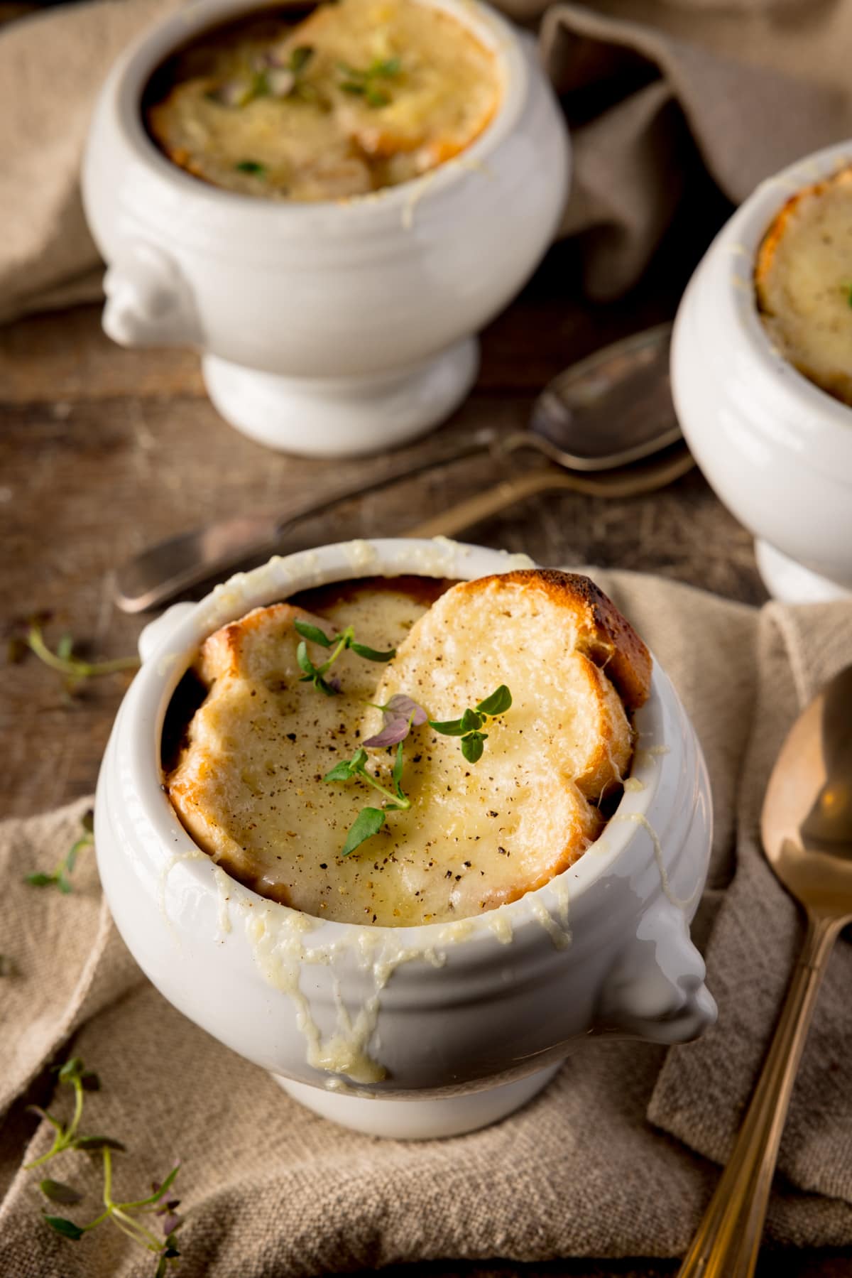 Three bowls of French Onion Soup on a wooden table. The soups are in white bowls and there is a beige napkin under the front bowl of soup. Each soup is topped with a little fresh thyme.