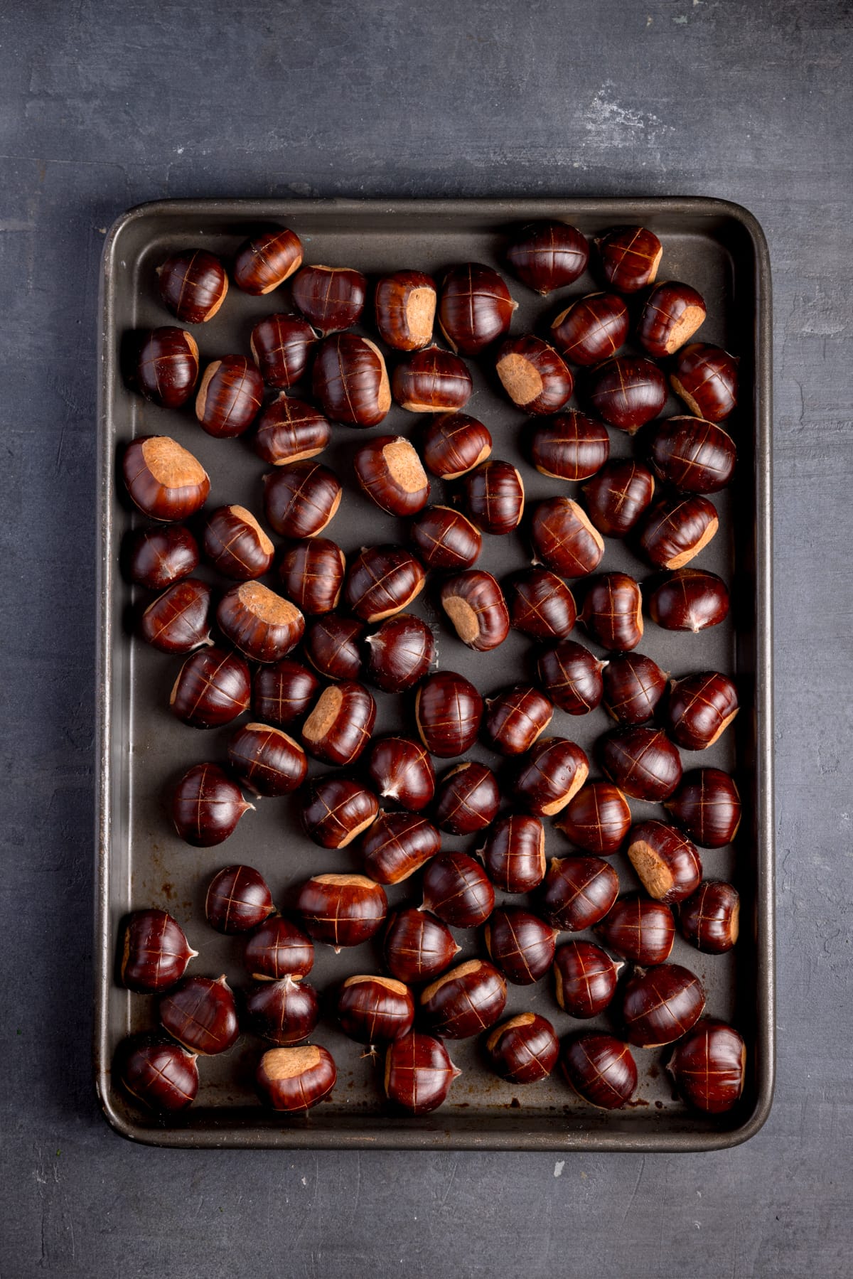 Raw chestnuts spread out on a metal baking tray with crosses cut in the top.