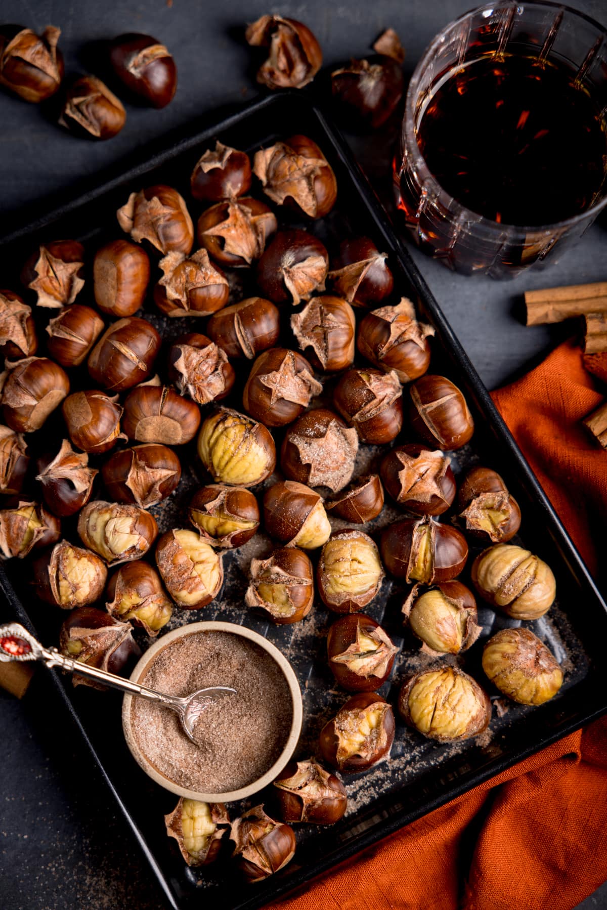 Picture of roast chestnuts on a dark metal tray with a small bowl of cinnamon sugar in the bottom left corner.