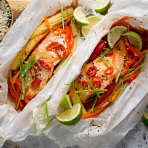 Square image of two salmon fillets cooked in individual baking parchment parcels with slices of chilli, peppers, babycorn, carrot and spring onion. Also topped with wedges of lime. The parcels are open, on a wooden board on a light background. There is a dish of sesame seeds nearby.