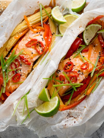 Square image of two salmon fillets cooked in individual baking parchment parcels with slices of chilli, peppers, babycorn, carrot and spring onion. Also topped with wedges of lime. The parcels are open, on a wooden board on a light background. There is a dish of sesame seeds nearby.