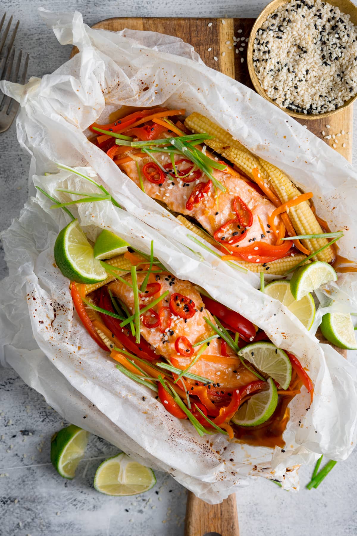 Overhead of two salmon fillets cooked in individual baking parchment parcels with slices of chilli, peppers, babycorn, carrot and spring onion. Also topped with wedges of lime. The parcels are open, on a wooden board on a light background. There is a dish of sesame seeds and some lime wedges nearby.