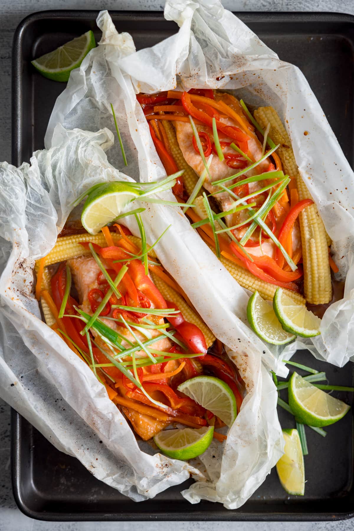 Two salmon fillets cooked in individual baking parchment parcels with slices of chilli, peppers, babycorn, carrot and spring onion. Also topped with wedges of lime. The parcels are open, on a dark baking tray. There are some lime wedges nearby.