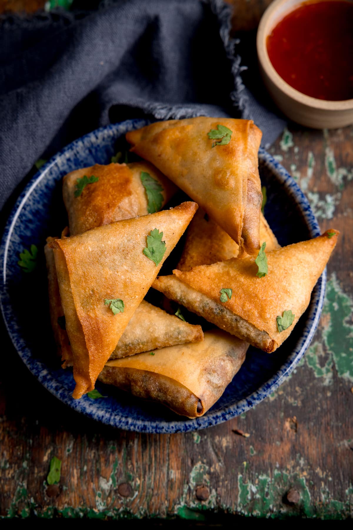 Samosas piled up on a small blue plate next to a grey napkin and little bowl of sweet chilli sauce. The place in on an aged wooden background.