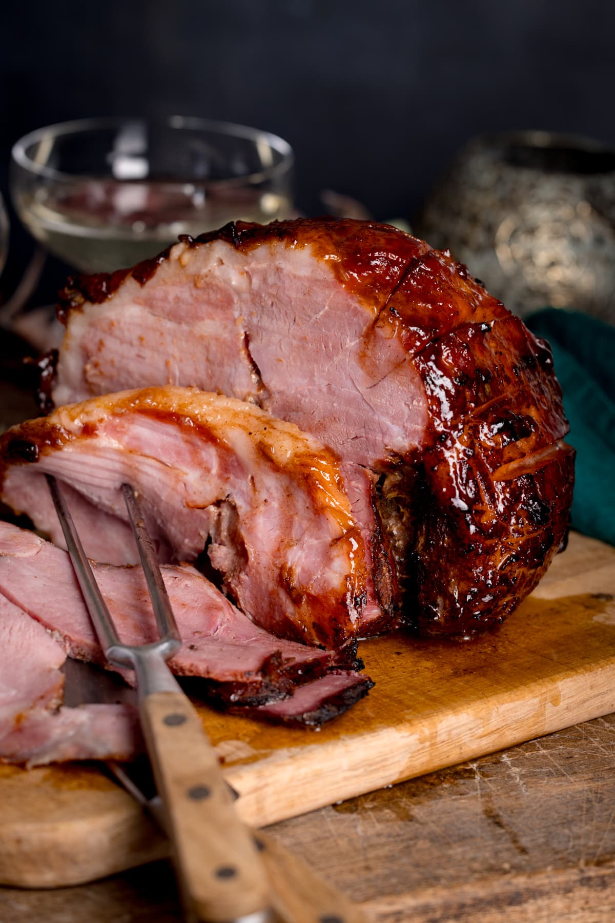 Close up picture of a slow cooker gammon joint with some slices sliced off and carving fork in the foreground all sat on a wooden chopping board.