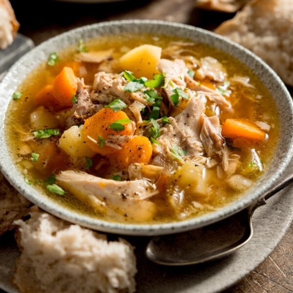 Square image of chicken and vegetable soup in a light bowl. The bowl is sat on a plate. There is a chunk of bread and a spoon nestled between the bowl and plate.