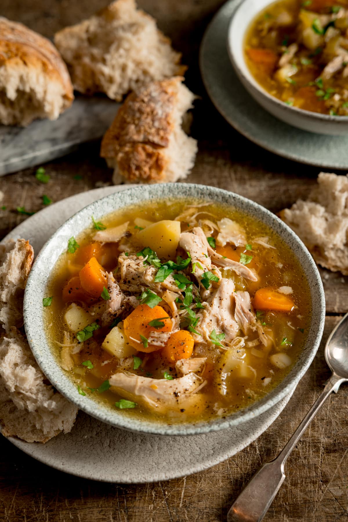 Tall image of chicken and vegetable soup in a light bowl. The bowl is sat on a plate. There is a chunk of bread nestled between the bowl and plate and there is a spoon next to the plate. The soup bowl is on a wooden table, and there is a further bowl of soup at the top right of the frame. There is a torn loaf of bread and some chunks of bread on a grey board on the top left of the frame.