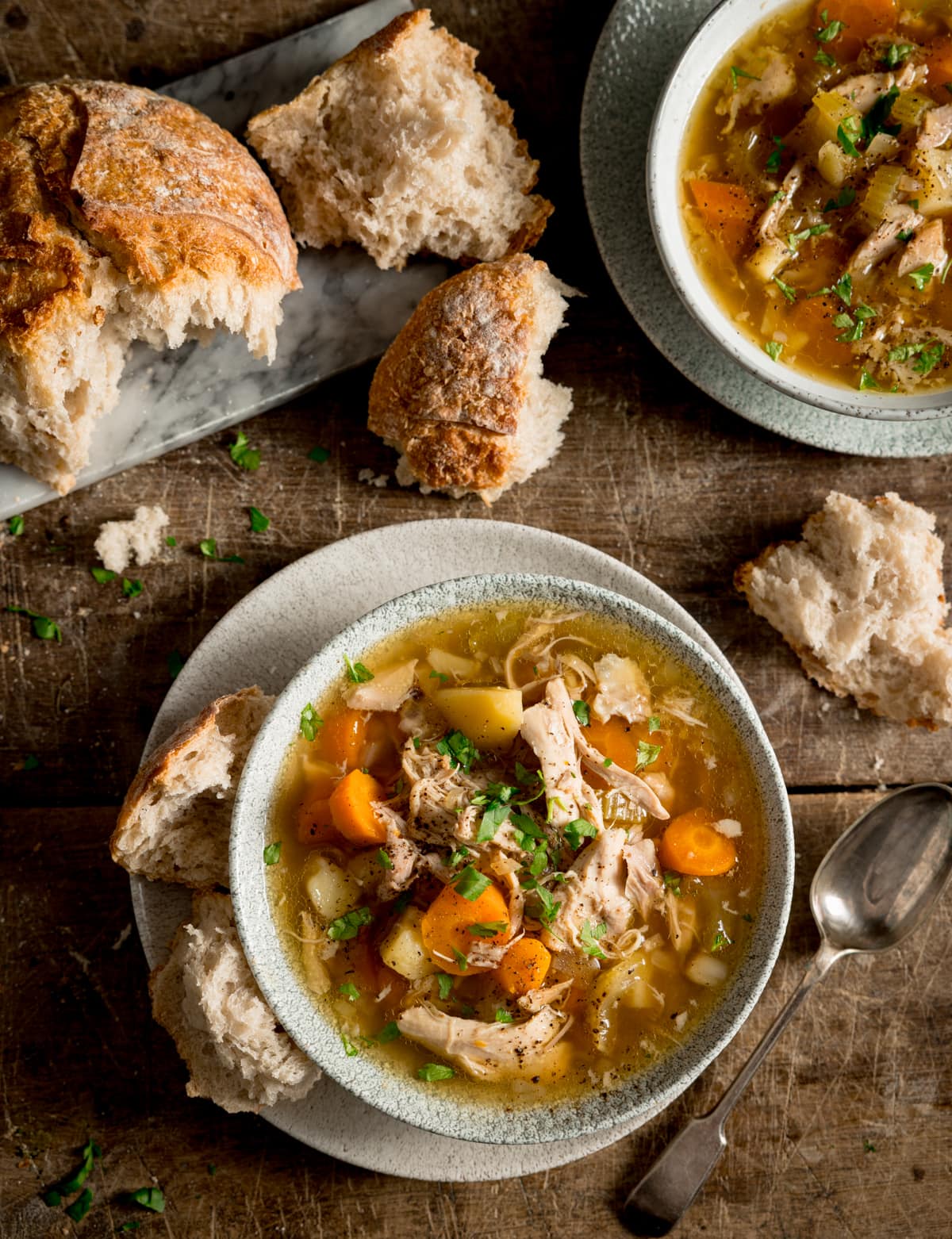 Tall overhead image of chicken and vegetable soup in a light bowl. The bowl is sat on a plate. There is a chunk of bread nestled between the bowl and plate and there is a spoon next to the plate. The soup bowl is on a wooden table, and there is a further bowl of soup at the top right of the frame. There is a torn loaf of bread and some chunks of bread on a grey board on the top left of the frame.