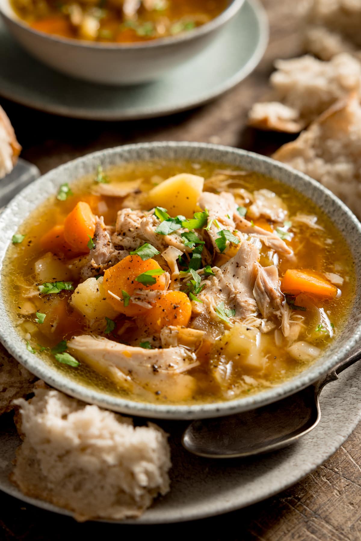Tall image of chicken and vegetable soup in a light bowl. The bowl is sat on a plate. There is a chunk of bread and a spoon nestled between the bowl and plate. The soup bowl is on a wooden table, and there is a further bowl in the background, along with some chunks of bread.
