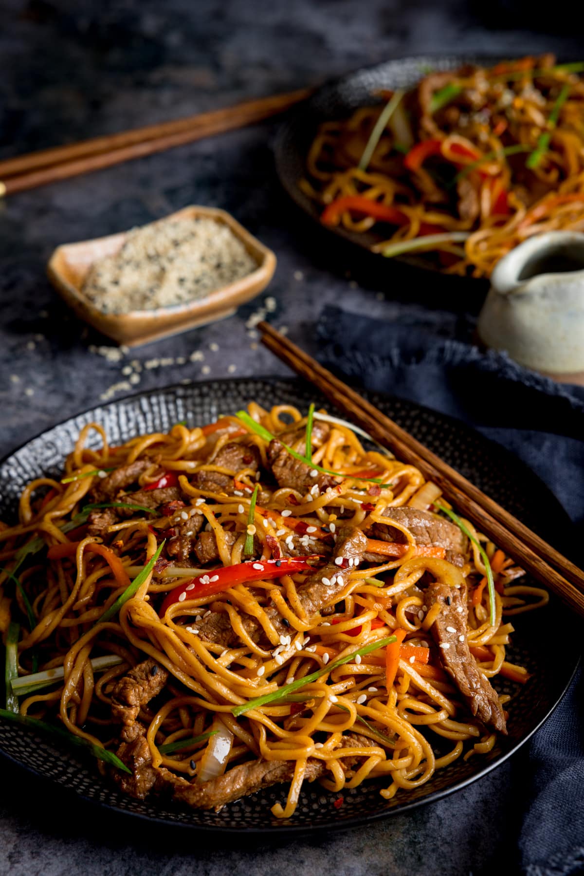 Tall image of beef teriyaki noodle stir fry on a black plate. There is a pair of wooden chopsticks resting on the plate. The plate is on a grey background. There is a little dish or sesame seeds and a further bowl of stir fry at the top o0f the frame.