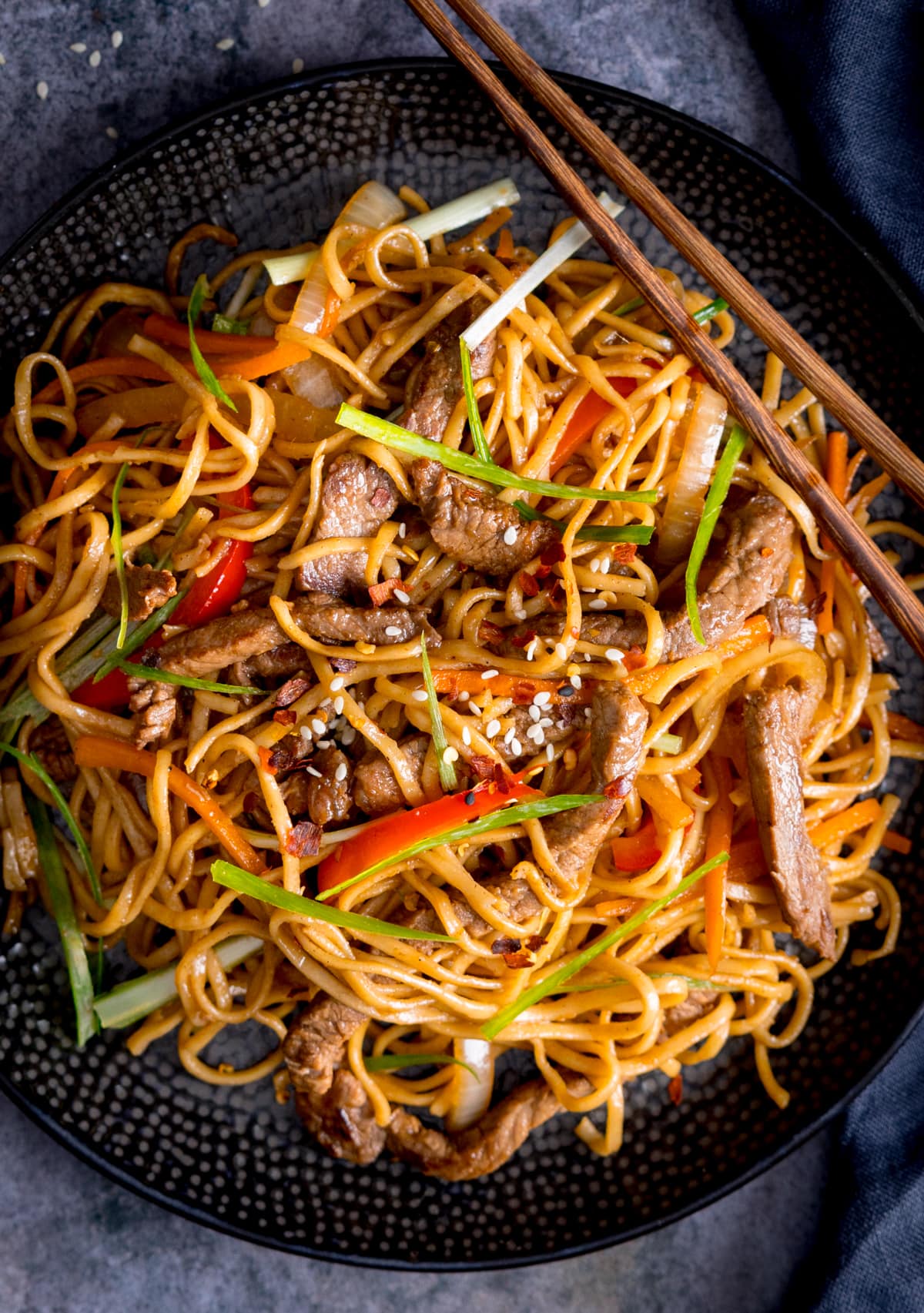 Tall overhead image of beef teriyaki noodle stir fry on a black plate. There is a pair of wooden chopsticks resting on the plate. The plate is on a grey background.