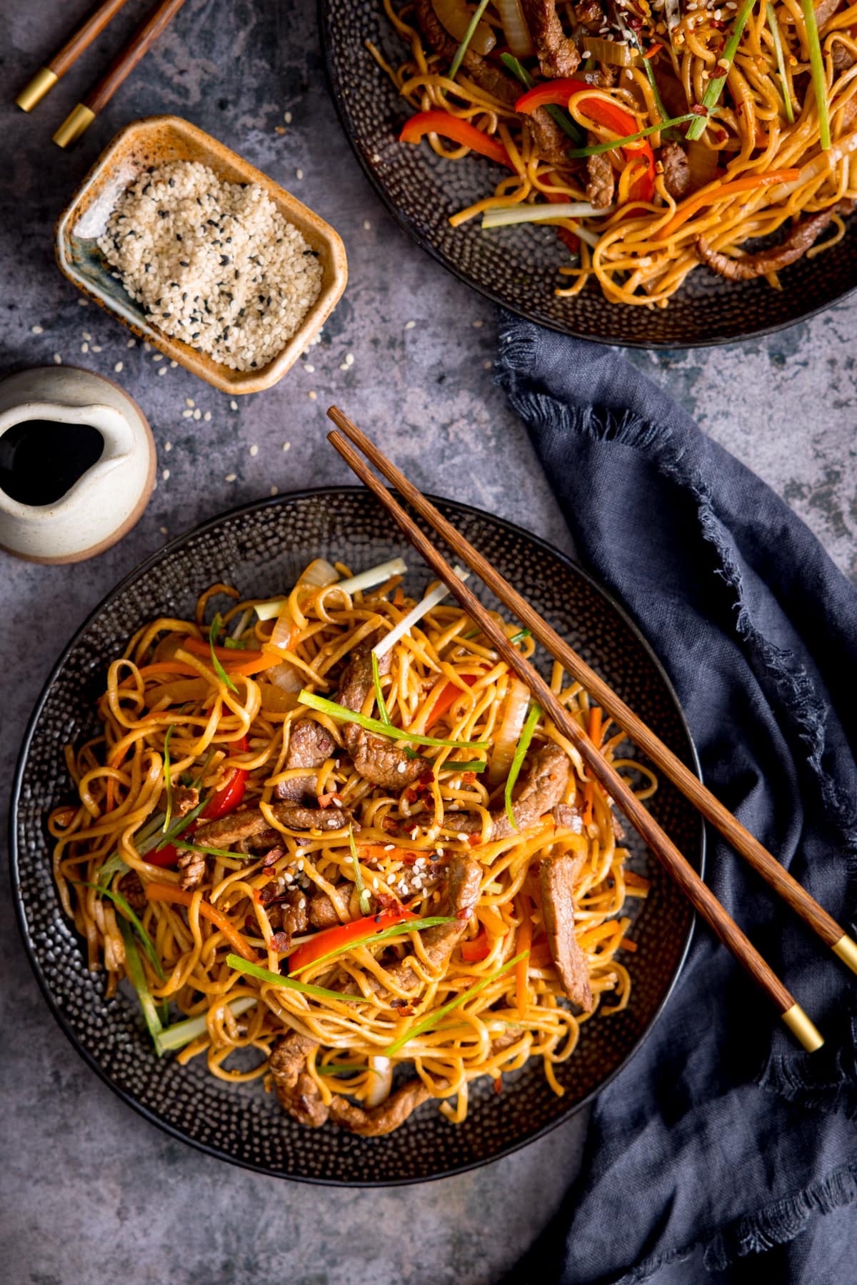 Tall overhead image of beef teriyaki noodle stir fry on a black plate. There is a pair of wooden chopsticks resting on the plate. The plate is on a grey background next to a navy napkin. There is a further plate with stir fry at the top of the frame. There is also a little jug of soy sauce and a little dish of sesame seeds on the top left of the main plate.