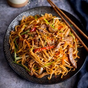 Square image of beef teriyaki noodle stir fry on a black plate. There is a pair of wooden chopsticks resting on the plate. The plate is on a grey background.