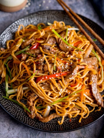 Square image of beef teriyaki noodle stir fry on a black plate. There is a pair of wooden chopsticks resting on the plate. The plate is on a grey background.