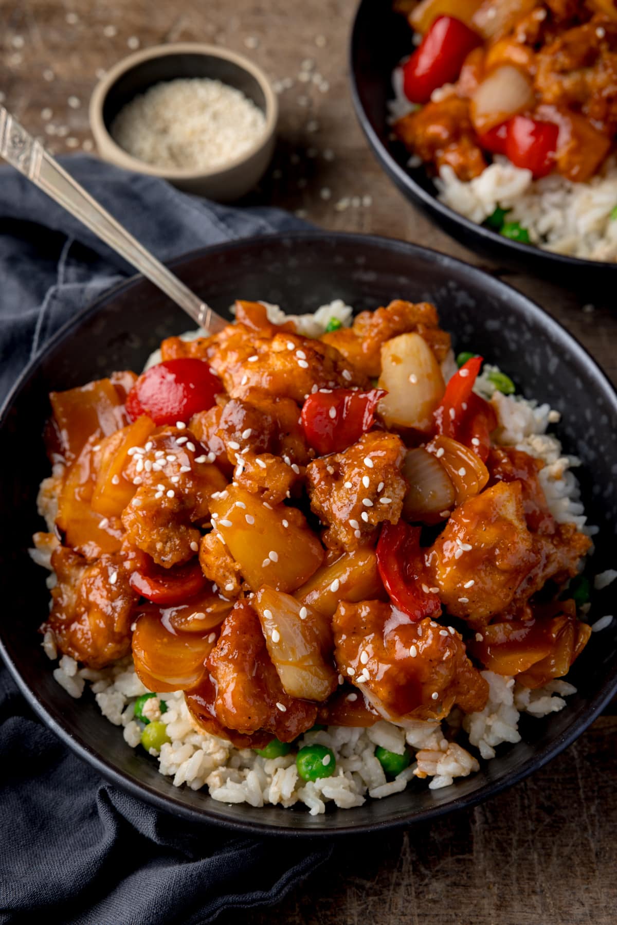 A dark bowl with baked sweet and sour chicken on a bed of egg fried rice with a fork sticking out of the top left corner.