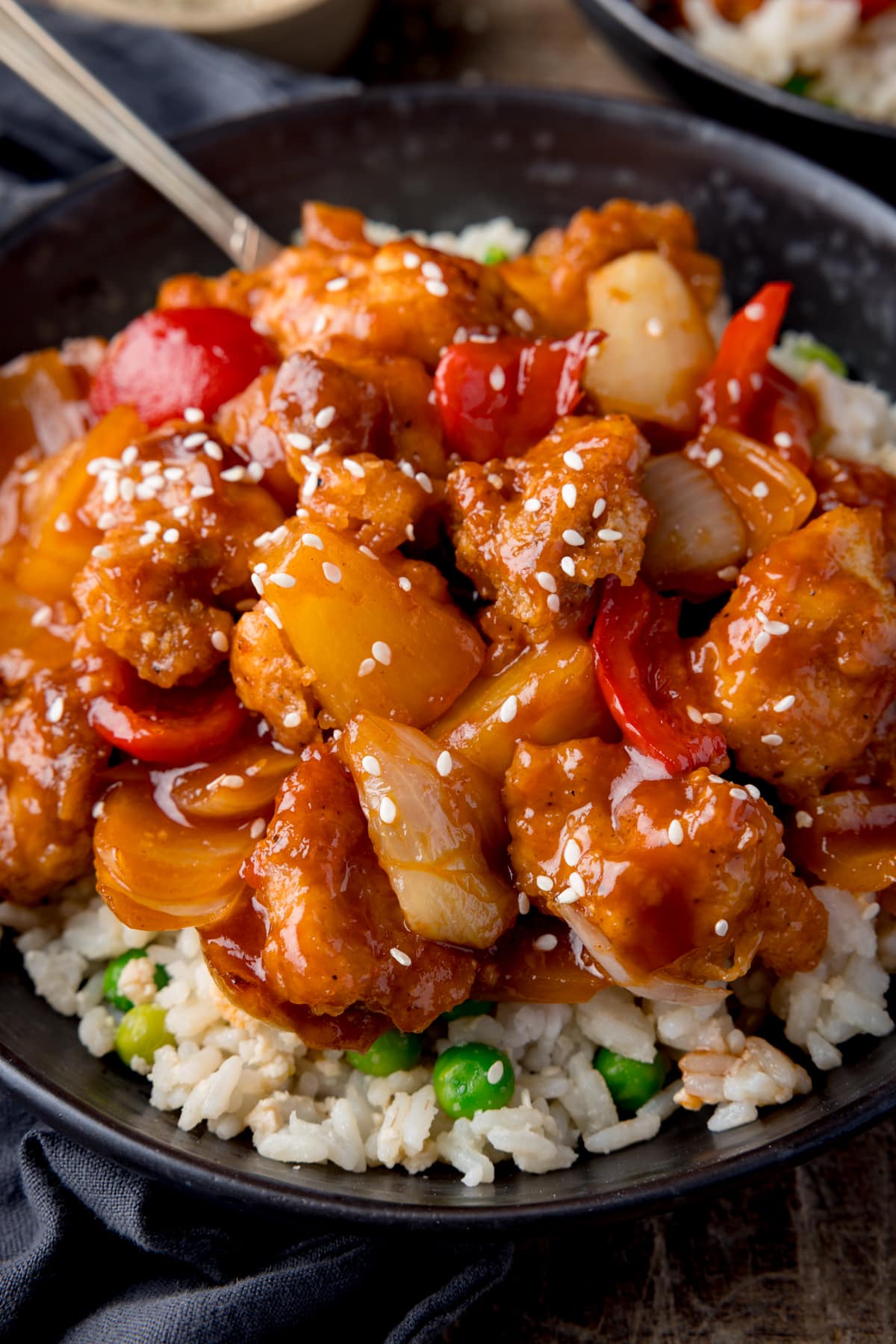 Close up picture of baked sweet and sour chicken on some egg fried rice in a dark bowl all sprinkled with light sesame seeds.