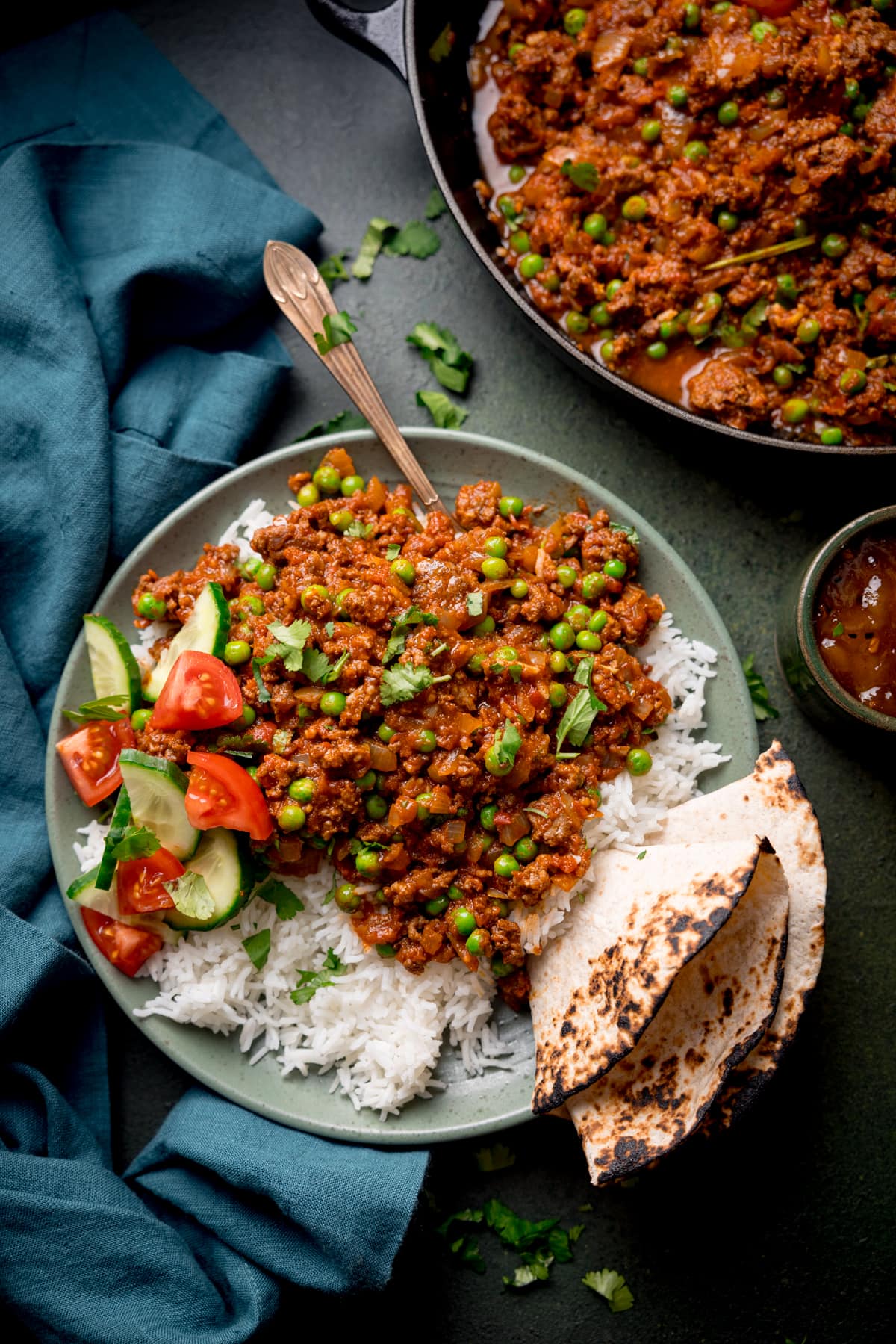 Overhead picture of a bowl full of Keema Curry sat on a bed of boiled rice rice with a chapati folded and wedged in the side of the dish and some sliced tomato and cucumber on the side.