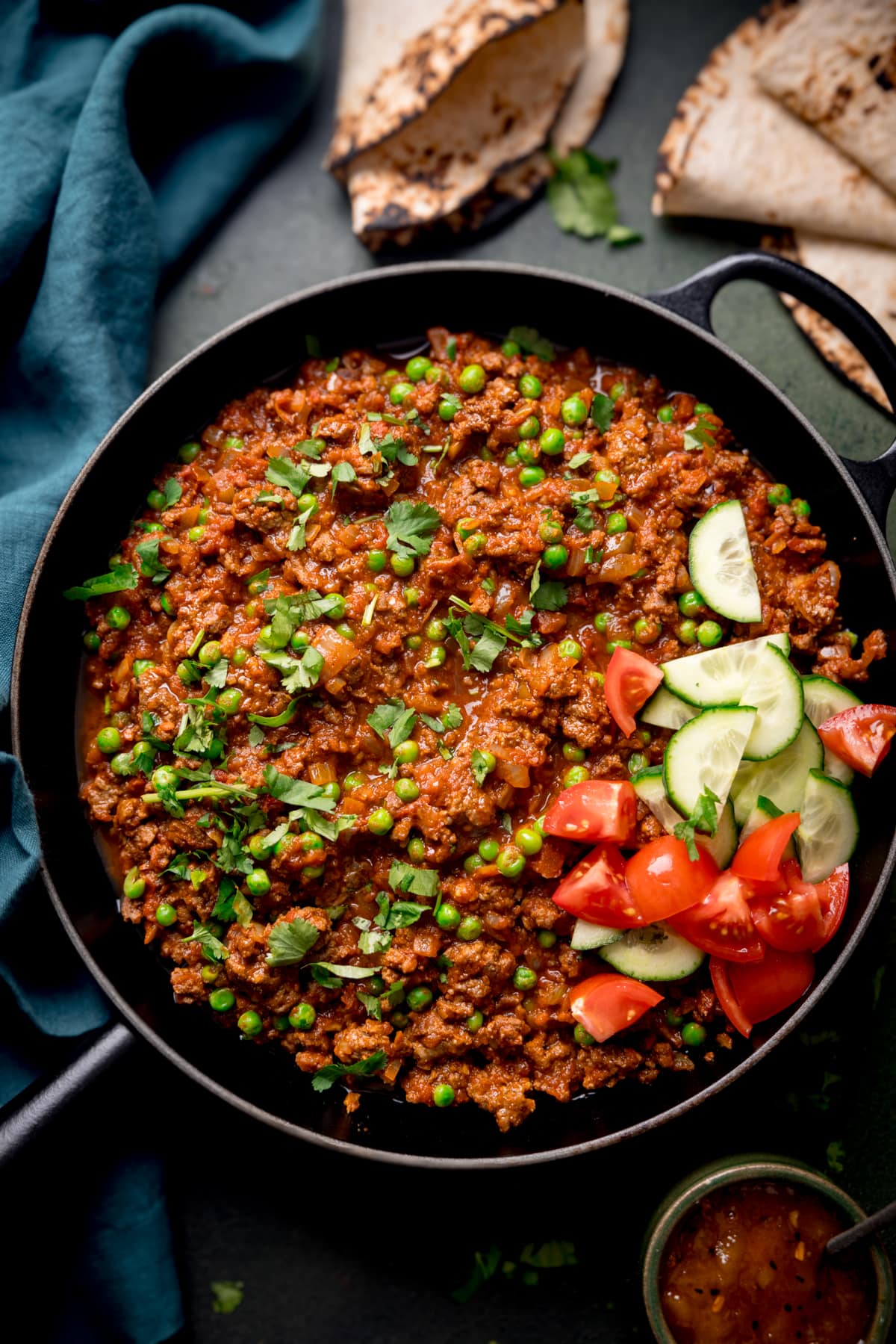 Overhead picture of a cast iron pan full of keema curry (minced beef curry) with some chapati and a dish full of mango chutney in the background.