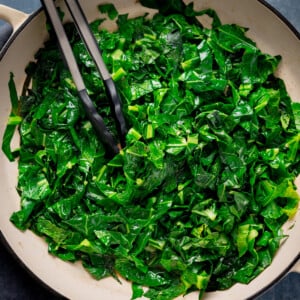 Overhead square image of pan fried spring greens in a white pan. There is a set of tongs nestled in the spring greens.