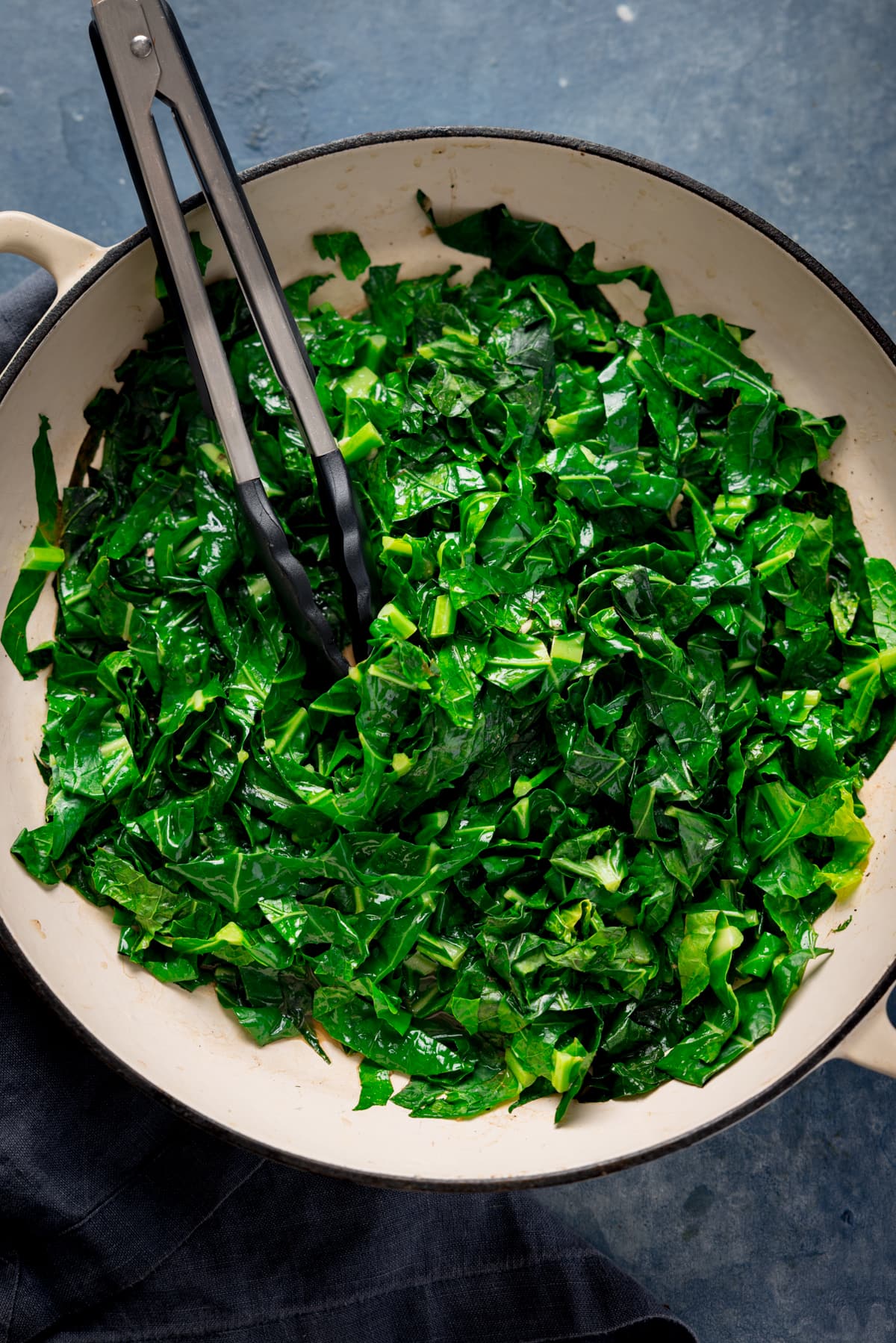 Overhead tall image of pan fried collard greens in a white pan. There is a set of tongs nestled in the spring greens. The pan is on a blue background.