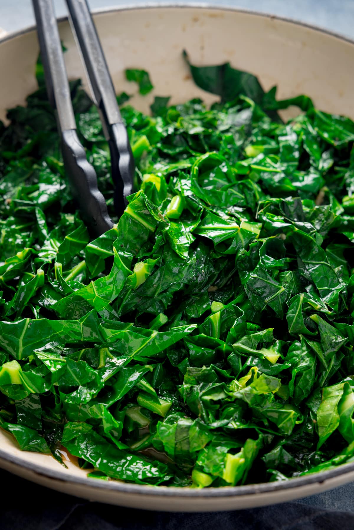 Close up tall image of pan fried spring greens in a white pan. There is a set of tongs nestled in the spring greens.