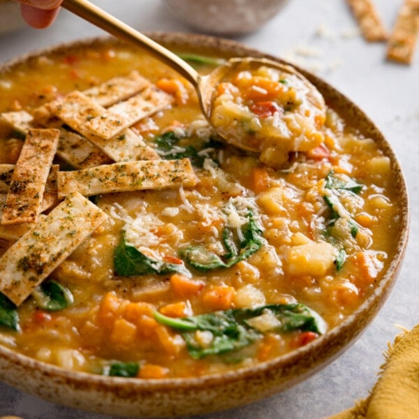 Red lentil soup topped with crispy tortilla strips in a brown speckled bowl. The bowl is on a light background. There is a gold spoon taking a spoonful of the soup.