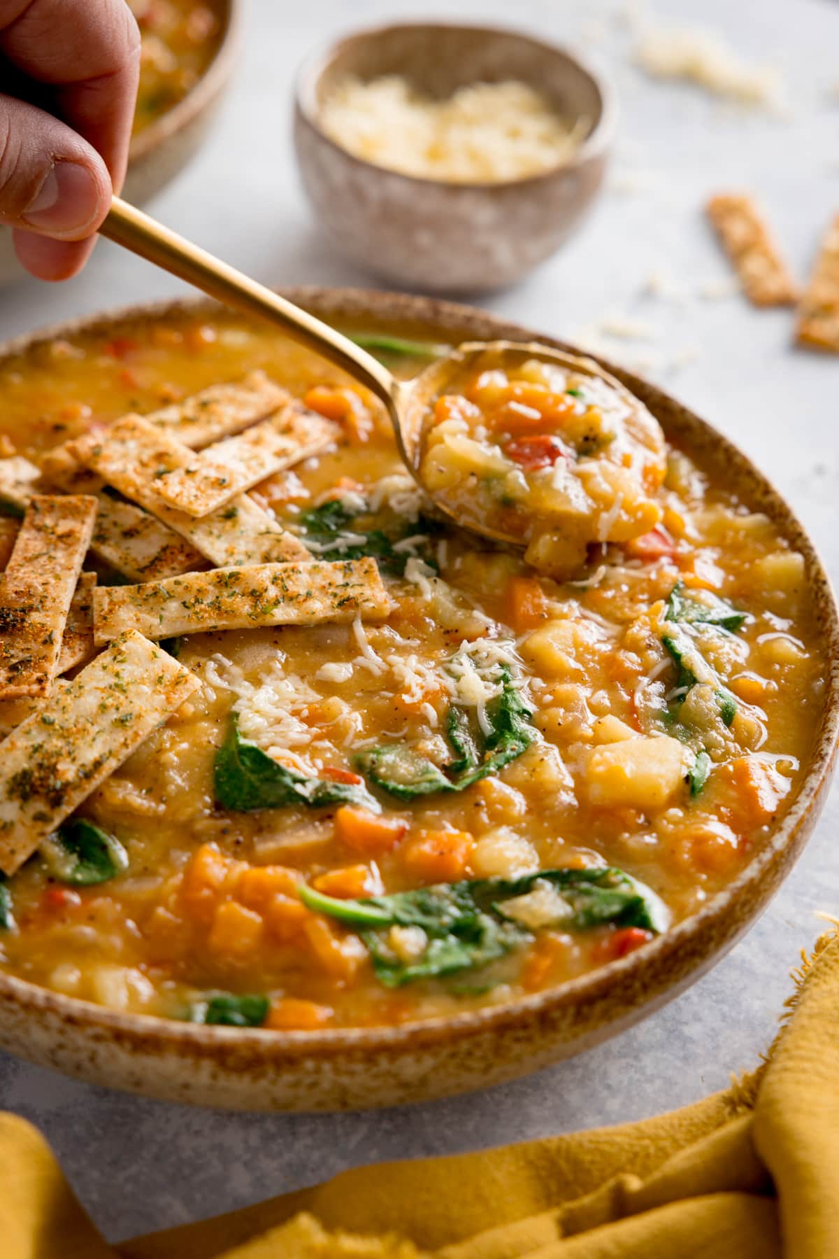 Tall image of red lentil soup topped with crispy tortilla strips in a brown speckled bowl. The bowl is on a light background. A spoonful of the soup is being lifted from the bowl. There are scattered tortilla strips and a small bowl of grated cheese at the top of the image and yellow napkin at the bottom of the image.