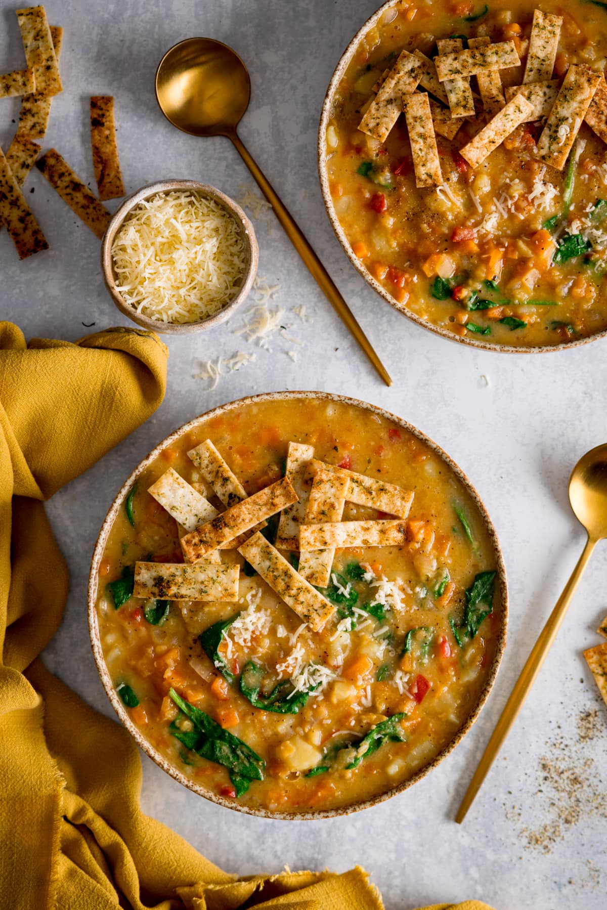 Tall image of two bowls of red lentil soup topped with crispy tortilla strips in brown speckled bowls. The bowls are on a light background. There is a yellow napkin on the left of the bottom bowl. There is a small bowl of grated cheese, two gold spoons and some scattered tortilla strips around the bowls.