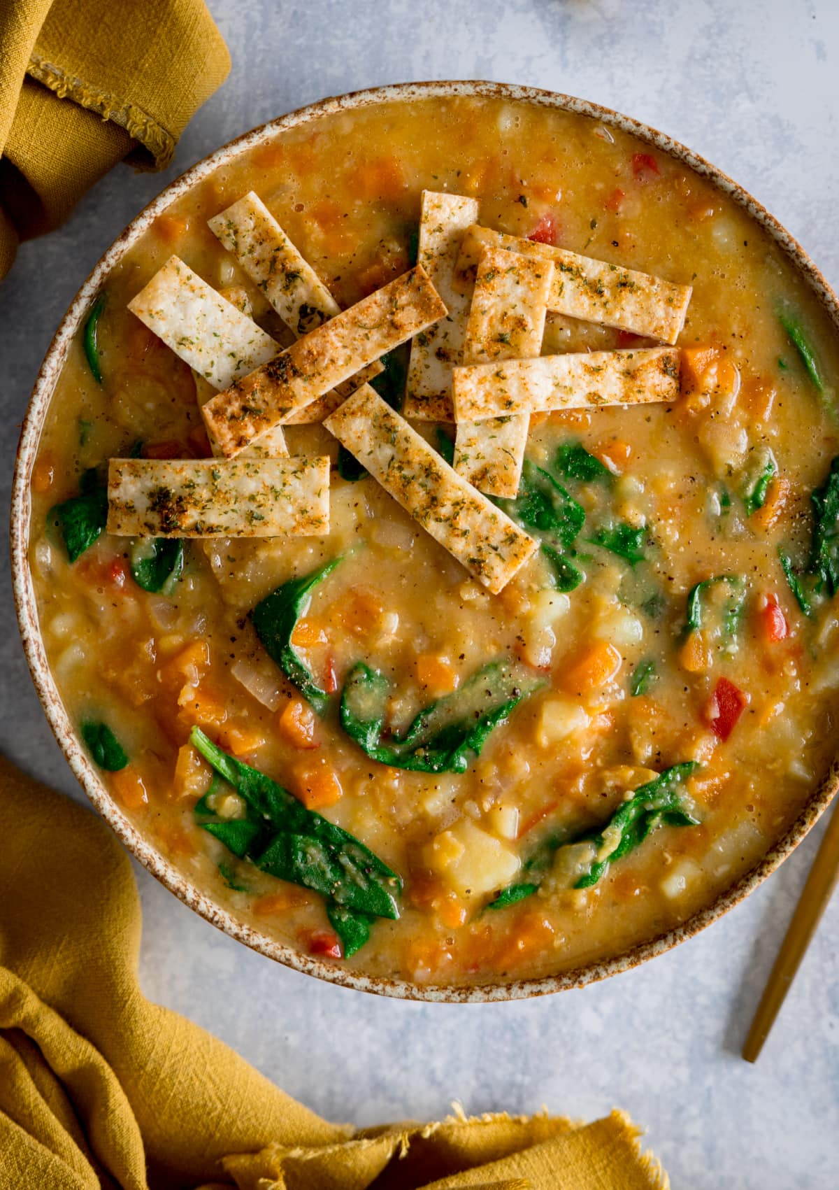 Tall image of red lentil soup topped with crispy tortilla strips in a brown speckled bowl. The bowl is on a light background. There is a yellow napkin on the left of the bowl.