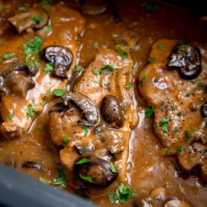 Square close-up image of smothered pork chops with mushrooms and gravy in the pan of a slow cooker. There is chopped parsley sprinkled on top.
