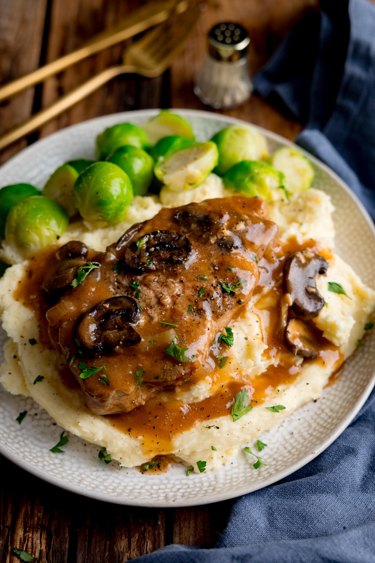 Slow cooker smothered pork chop with gravy and mushrooms on top of a pile of mashed potato, next to sprouts on a white plate. The plate is on a wooden background next to a blue napkin. There is gold cutlery and a small salt shaker at the top of the image.