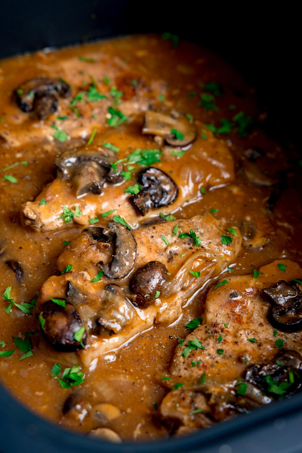 Tall close-up image of smothered pork chops with mushrooms and gravy in the pan of a slow cooker. There is chopped parsley sprinkled on top.