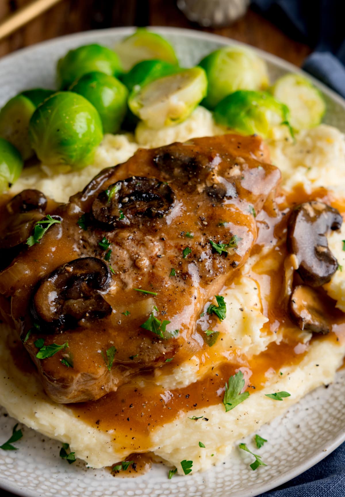 Slow cooker smothered pork chop with gravy and mushrooms on top of a pile of mashed potato, next to sprouts on a white plate. The plate is on a wooden background next to a blue napkin. There is some gold cutlery at the top of the image.