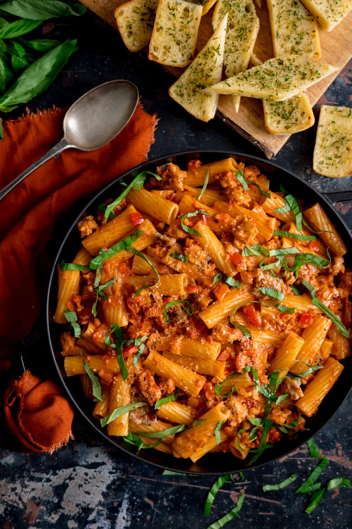 Overhead photo of a black frying pan full of spicy sausage rigatoni with some garlic bread, a rustic spoon and some fresh basil on the side.