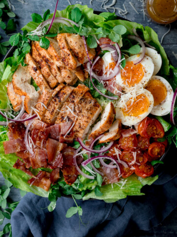 Overhead square image of a large BLT chicken salad with soft boiled eggs in a bowl on a blue background. The bowls is next to a wooden board, a blue napkin and a little bottle of salad dressing at the top of the frame.