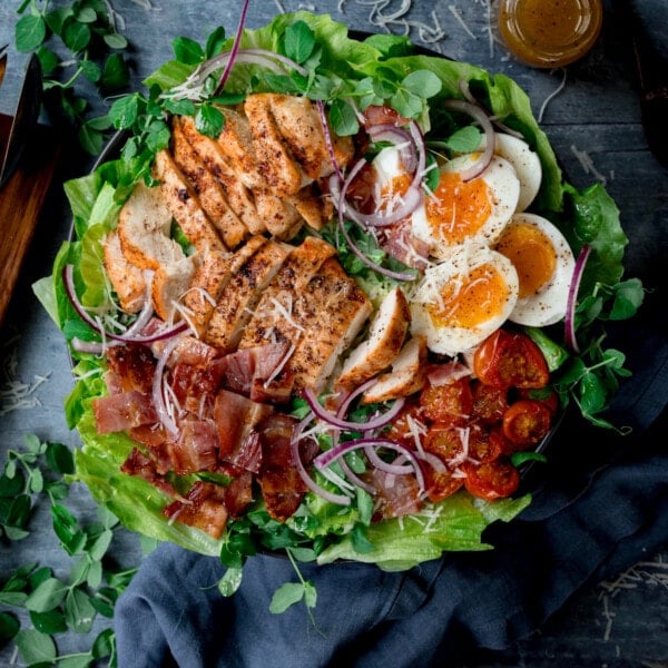 Overhead square image of a large BLT chicken salad with soft boiled eggs in a bowl on a blue background. The bowls is next to a wooden board, a blue napkin and a little bottle of salad dressing at the top of the frame.