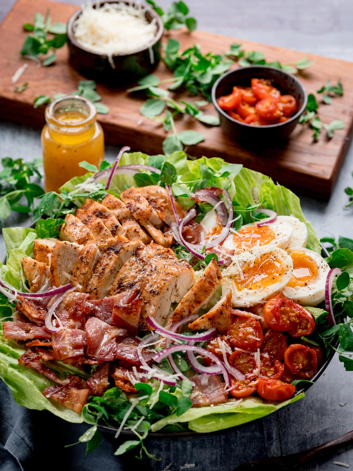 Tall image of a large BLT chicken salad with soft boiled eggs in a bowl on a blue background. There is a wooden board in the background with extra ingredients on it (tomatoes, parmesan and pea shoots). There is also a little bottle of salad dressing next to bowl.