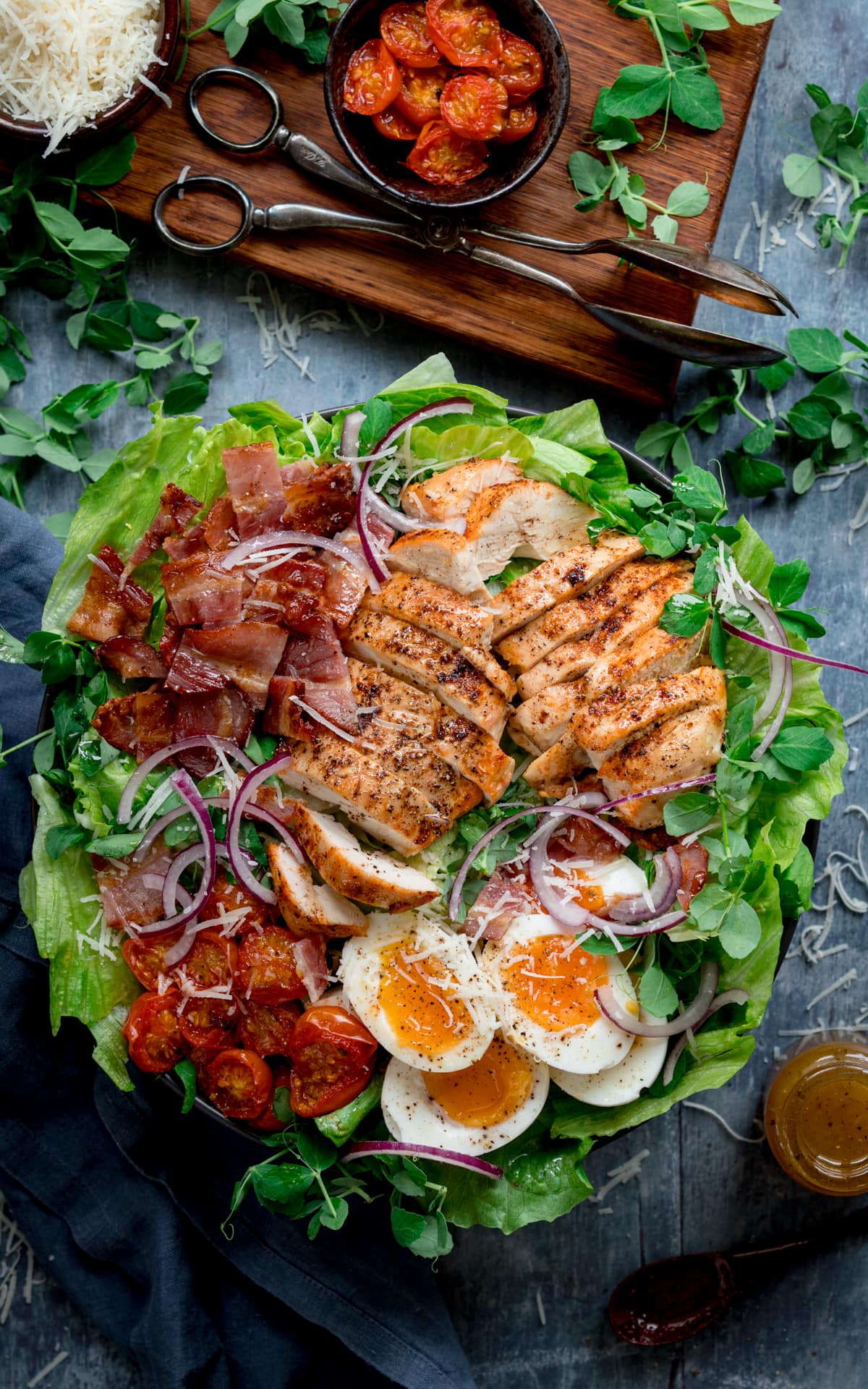 Overhead tall image of a large BLT chicken salad with soft boiled eggs in a bowl on a blue background. The bowls is next to a wooden board that has extra ingredients on it (tomatoes, parmesan and pea shoots), plus some salad servers. There is also a little bottle of salad dressing to the right of the bowl.