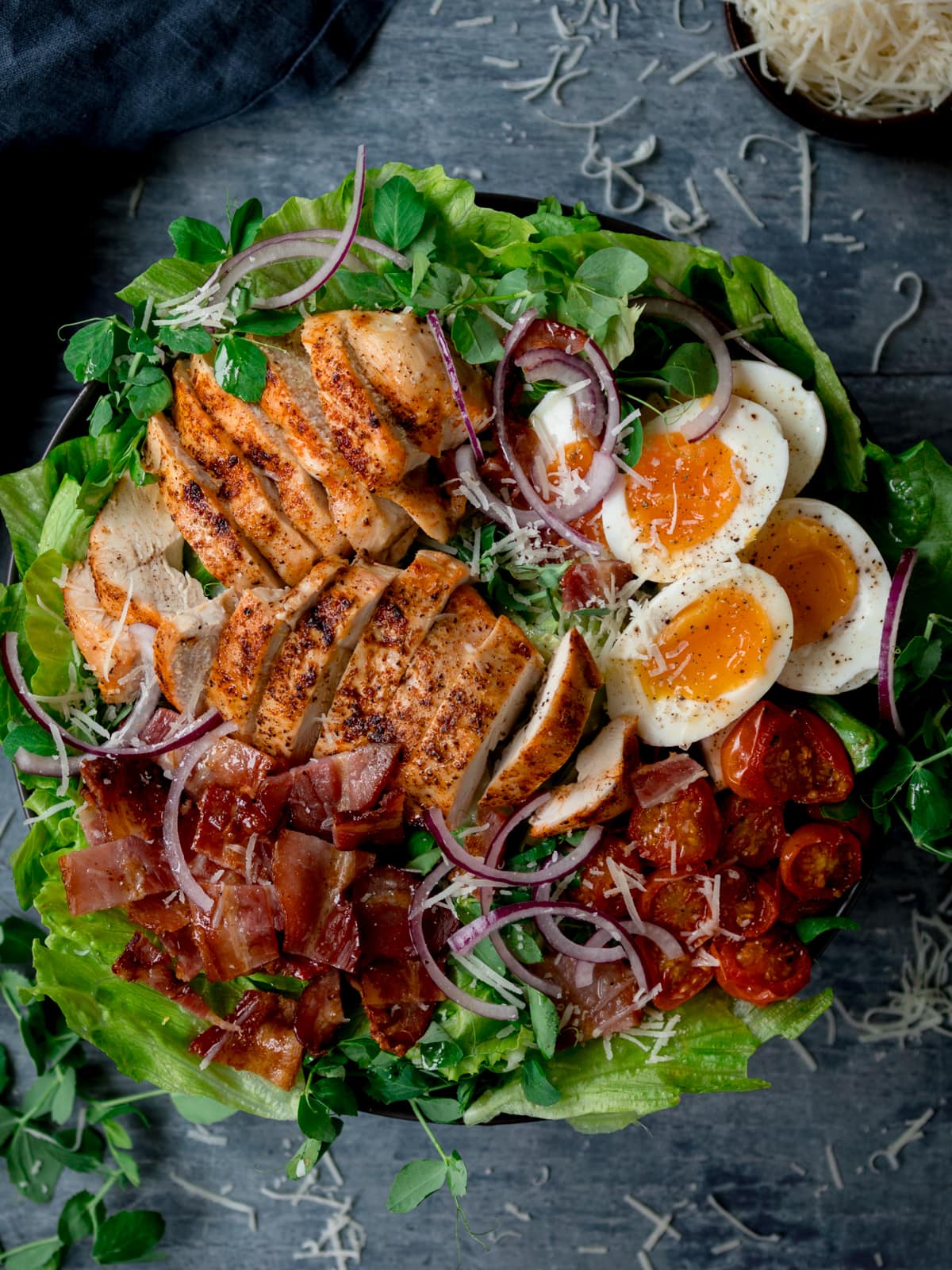 Overhead tall image of a large BLT chicken salad with soft boiled eggs in a bowl on a blue background.
