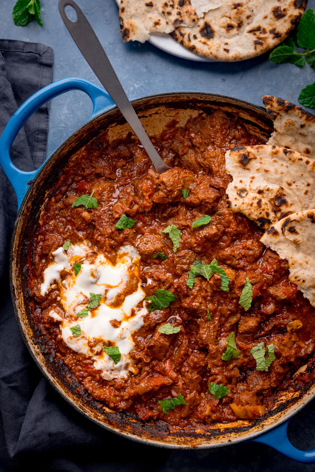 Overhead tall image of Beef Rogan Josh in a blue pan. The curry is topped with chopped mint leaves, a swirl of plain yogurt and some torn chapati.