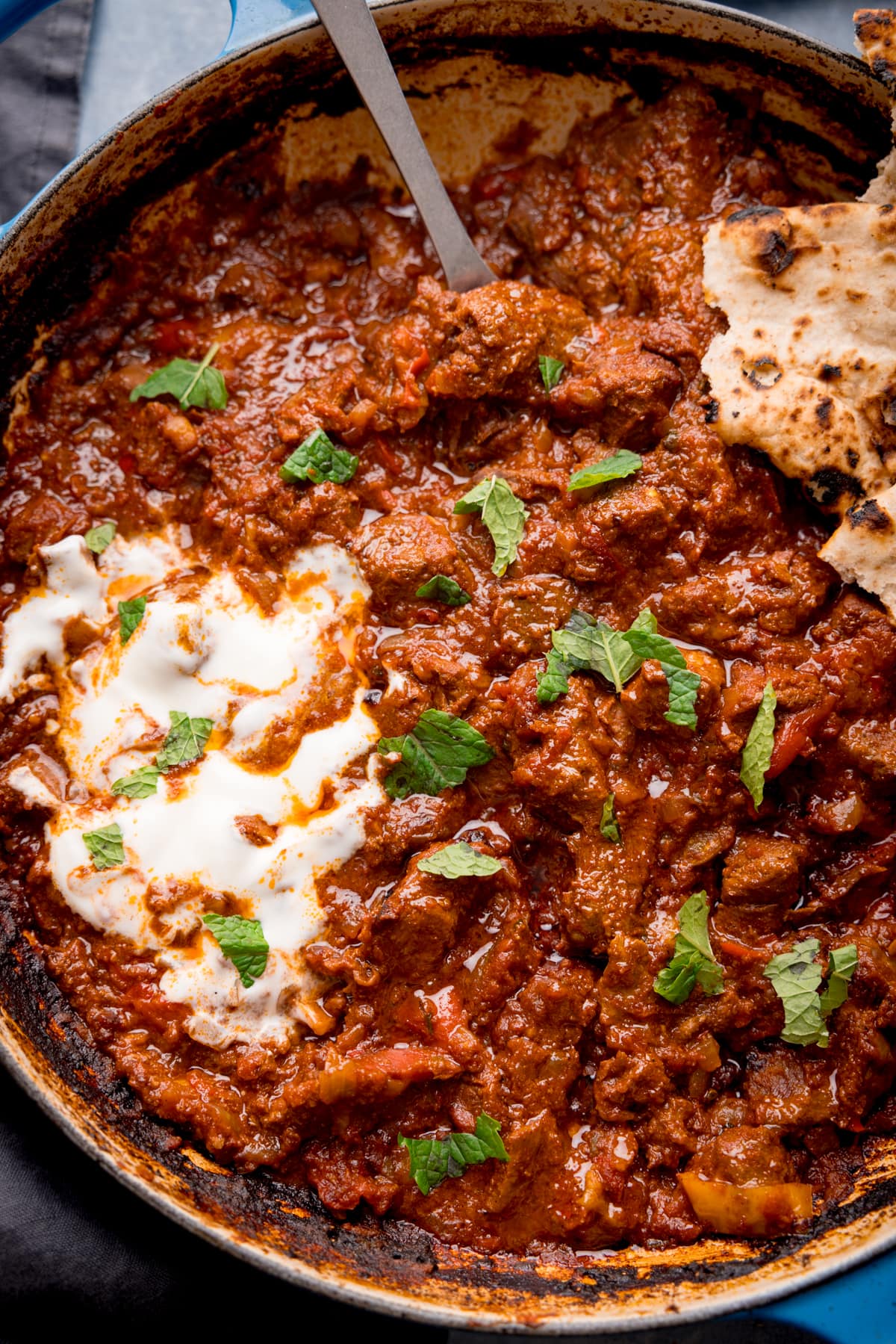 Overhead close-up image of Beef Rogan Josh in a blue pan. The curry is topped with chopped mint leaves, a swirl of plain yogurt and some torn chapati. There is a silver serving spoon sticking out of the curry.